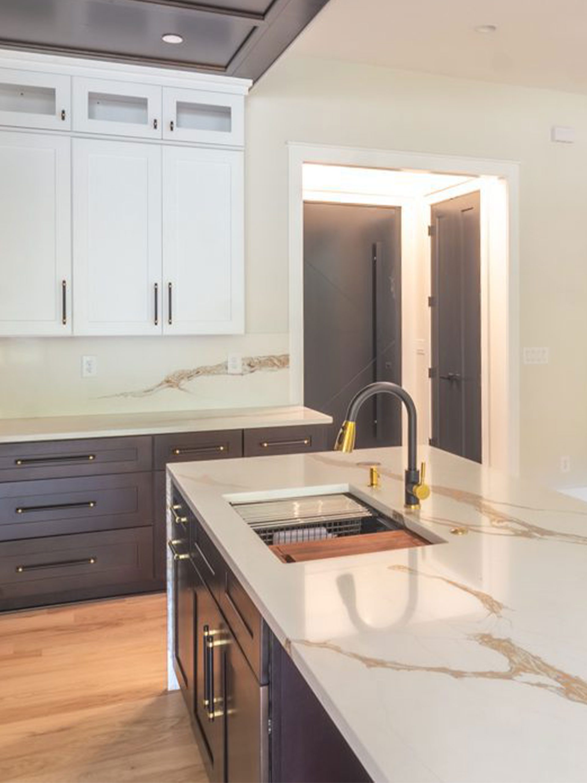 Modern kitchen with a large island, gold faucet, marble countertops, dark lower cabinets, white upper cabinets, and light wood flooring.